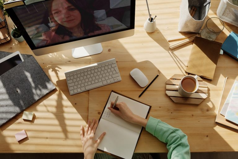 Person taking notes during a video call on a computer, with various office supplies and a cup of coffee on the desk.