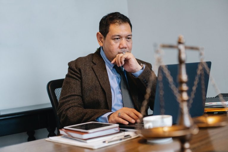 Man in a brown suit working on a laptop with a scale of justice in the foreground.
