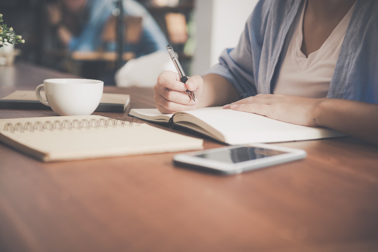 Personne écrivant dans un carnet avec une tasse de café et un smartphone sur la table.