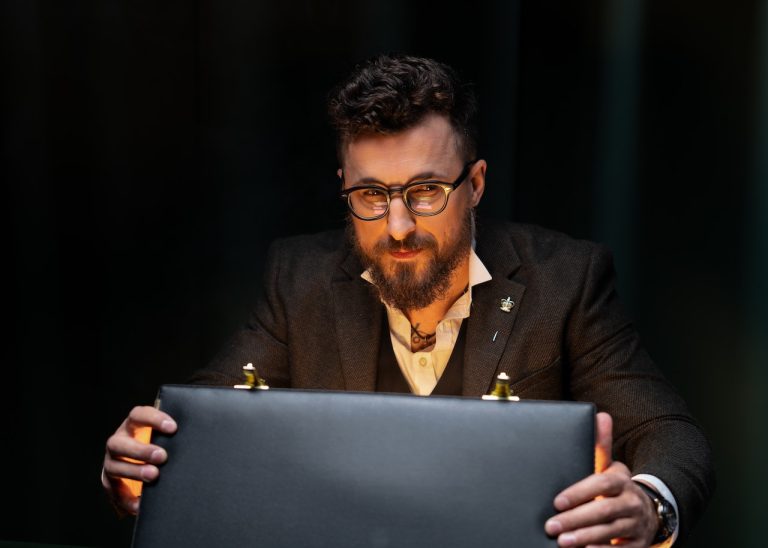 A man with glasses and a beard is sitting at a desk, wearing a suit and holding a briefcase, suggesting the seriousness and professionalism of a private investigator.