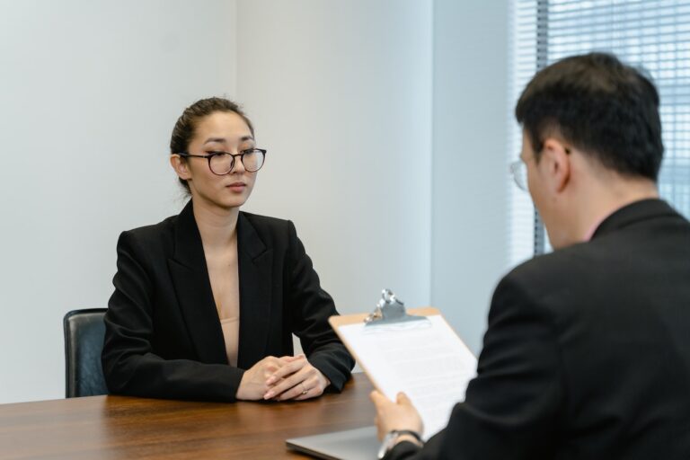 A woman in glasses sits at a table, engaged in a serious discussion with a man holding a clipboard. They are in a professional setting, indicating an interview or a forensic interrogation scenario.